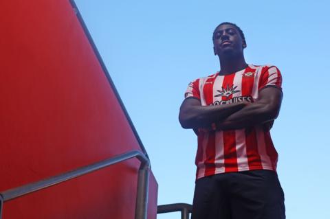 Divin Mubama looks down at the camera with his arms crossed while wearing Southampton's red and white home kit
