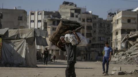 A man with grey hair carries belongings on his shoulders as he walks through a Gaza City street littered with debris and rubble.