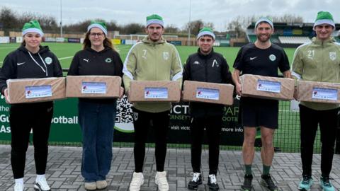 Two women and four men  dressed in sports kit are standing on front of a football pitch. They are all wearing green santa hats and holding boxes wrapped in Christmas paper, with greetings tags on them.