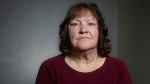 A woman sitting against a softly lit, neutral background, looking toward the camera with a calm, serious expression and wearing a burgundy top.