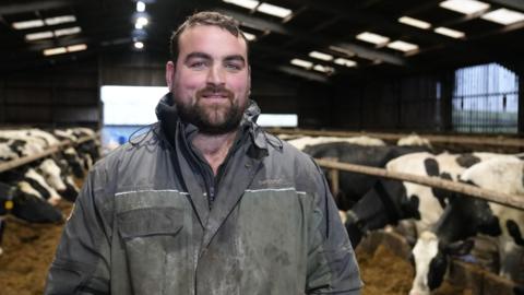 Gethin Hughes, dairy farmer stands in his cattle shed wearing blue overalls