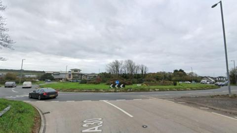 An approach to a roundabout from a dual carriageway To the left in the background is a retail complex.