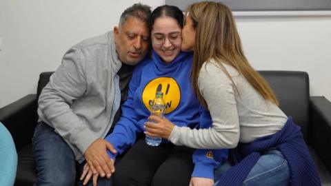  Liri Albag wearing a blue hoodie with a yellow smiley face sits in between her parents who are embracing and kissing her.