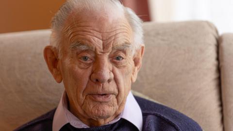 A close up of Barry. He has short grey hair and is wearing a navy jumper, with a pink shirt underneath.