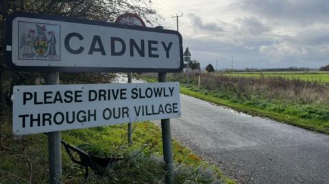 The picture shows a village sign on the left hand side, with the words 'Cadney' and 'please drive slowly through our village' written on it. To the right hand side is a road, and fields.