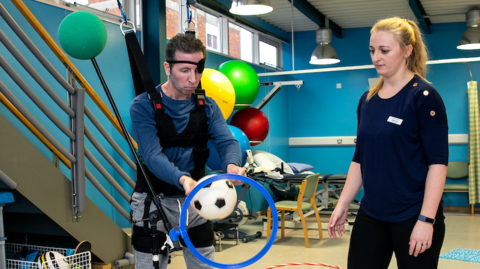 A man wearing a harness around his chest and legs holds a football in his hand and attempts to pass it through a hoop at waist height. He is watched by a woman in dark clothing who has a name badge on her chest that is not readable. 