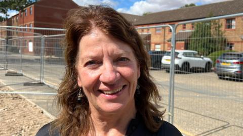 A woman with shoulder-length brown hair and wearing silver earrings. She is standing in front of a silver barrier which is in front of a car park located outside a two-storey building.