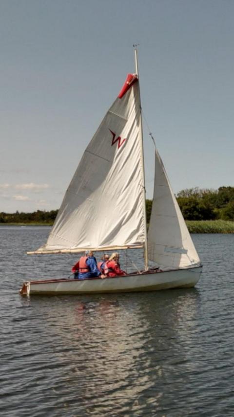 A small white sailboat with two large sails is in the middle of a calm lake. Three people wearing red life jackets sit in the boat, whilst trees line the background. 