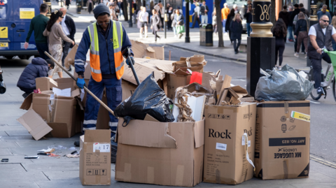 A refuse worker sweeps around a pile of cardboard boxes left on the pavement on Oxford Street
