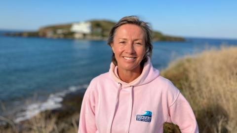 A woman in a pink hoody stands on grassland above a beach. It is sunny and you can see the sea and part of the beach behind her. 
