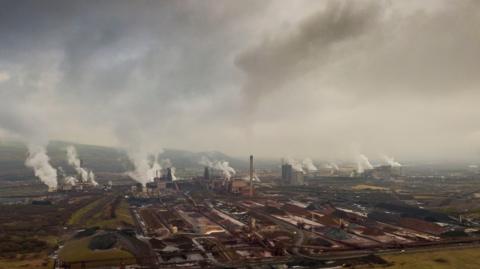 A panoramic view of the huge Port Talbot steelworks, showing numerous plumes of smoke from various structures against a grey sky