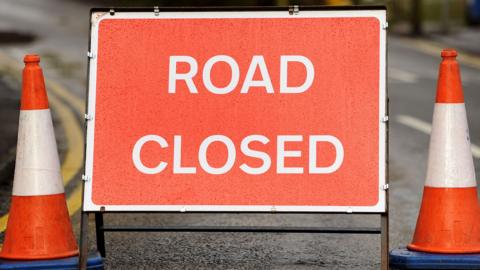 A picture of a red sign with the words 'ROAD CLOSED' in white. There are two orange and white traffic cones either side of it.