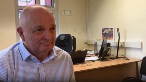 A man with a bald head, wearing a blue and white striped shirt, chats to a reporter off-camera in his office. This is a still taken from a TV interview. 