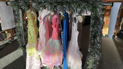 Brightly-coloured costumes, including pink and yellow dresses, hanging on a tinsel-covered rail. There is a shelf with books on it to the left of the image.