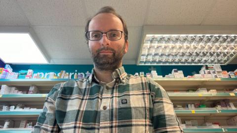 Man looking down at the camera. He's in a pharmacy with boxes of medicine in the background. He's got glasses on and dark black hair. He's wearing a green, stripy shirt.