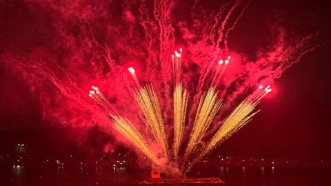 Gold and red fan shaped firework in fan shape, over water.