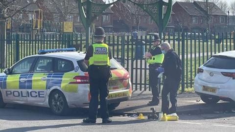 Three police officers stood near evidence markers and a police car