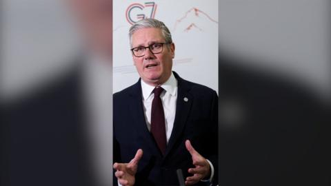 Keir Starmer standing in front of a G7 logo gestures with his hands while speaking to a journalist.
