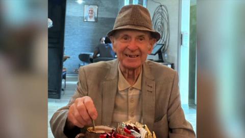 An elderly man smiles (right) for a photo in a cafe. He is wearing a pork-pie hat and a cream jacket and light shirt. He is stirring a hot drink in a mug on a table in front of him which contains a mental container of condiments