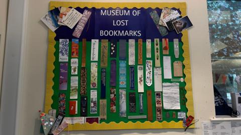 Around 50 bookmarks are stuck to a green display board. They are all different - some are classic bookmark shaped but others are postcards, tickets, children's artwork, or notes on torn pieces of paper.