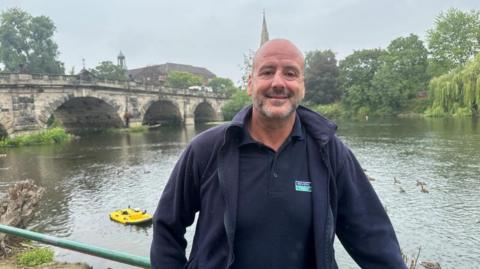 A bald man standing in front of the River Severn. A large stone bridge with four archways his behind him in the distance as well as a church spire. The man is wearing a navy blue polo shirt and a navy fleece. He is smiling and leaning on a green railing. There is a bright yellow paddle board on the river behind him to the left. 