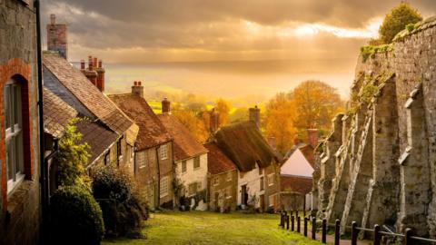 Gold Hill in Shaftesbury. A row of houses on a steep hill. There is a large stone wall on one side. The sky is glowing orange with fields and hills behind.
