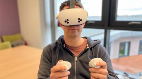A man wearing a virtual reality headset and holding controllers out in front of him. He is sitting in a room in front of a table with a window behind him.