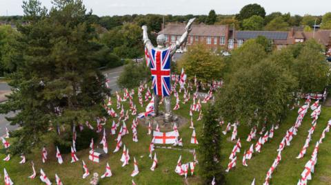 An aerial view shows the silver miner statue draped in a large Union Jack flag, its arms outstretched above rows of St George's flags covering the grass. Mature trees partially frame the statue on both sides. Brownhills Community Centre and surrounding roads are visible in the background.