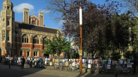 A lamp on a street in Cambridge. There are railing at the back covered in posters and bicycles locked to the railings. There is a church to the left