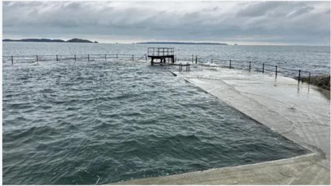 The diving board is pictured at high tide against a grey sea and grey sky.
