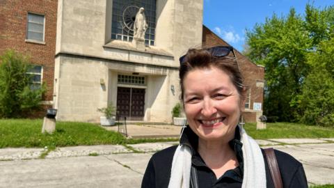 Mary Simons smiling outside of a stone fronted church with weeds in the background.