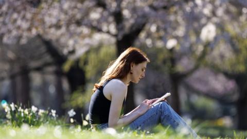 A woman uses her mobile phone while sitting on the grass in a sunny park, with trees in the background