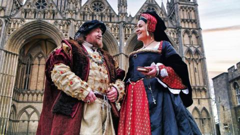 Two performance artists dressed in costumes from the Tudor era. A man is wearing a white long dress and a red gown with a black velvet hat. The woman beside him is wearing a black and red dress and a headpiece. They are both smiling at each other and standing in front of a cathedral. 