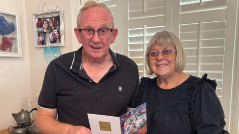Keith and Helen pictured standing in their home. He wears a black polo shirt and she wears a navy blouse. Both smile - Keith holds a Christmas card. Both have grey hair and wear glasses.
