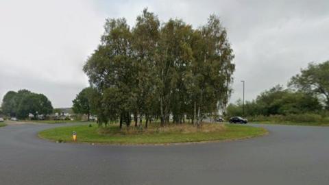 A roundabout with a cluster of tall, leafy trees planted in the centre on a grassy island. The road surrounding the roundabout is wide and paved, with a smooth, dark surface. Several streetlights are positioned around the roundabout, and a few vehicles can be seen in the background, including a black car approaching from the right side. The surrounding area appears to be open and green, with additional trees and shrubs visible beyond the roundabout. The sky is overcast.