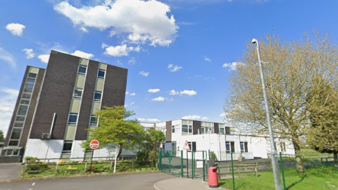 Two 1960s school buildings behind fences. One building is four storeys, the other two. Both are a mixture of white and dark grey concrete.