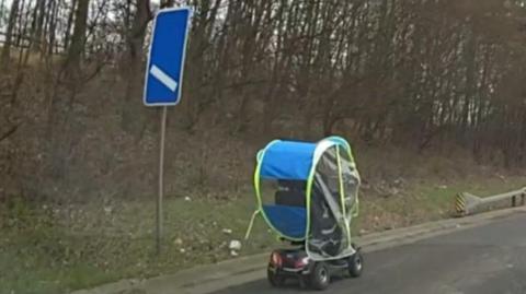 A mobility scooter with a rain cover drives along the hard shoulder. There are trees along the bank, with no leaves on them. There is a blue motorway sign to the left of the frame.