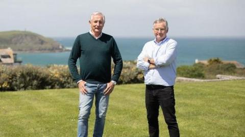 Two men wearing jeans and shirts standing on a lawn in the sunshine against a backdrop of blue sea and picturesque cliffs.