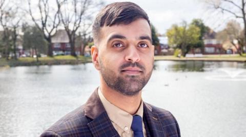 A man with short black hair and a short beard, wearing a suit jacket, shirt and blue tie, stands looking at the camera. A lake is behind him with trees and houses visible on the other side.