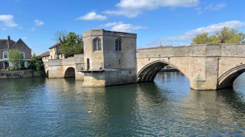A medieval bridge with limestone arches runs across a river with a small building overhanging one side, which is a chapel.