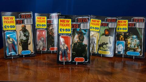A mixture of five Star Wars figurines, all standing on a wooden table. all with £1.59 price tags on them. They are all in their original boxes. 
