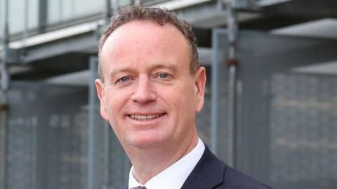 Stephen Kelly pictured in a tie and suit standing at the Ulster University campus in Londonderry.