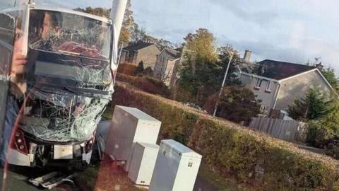 Image of a double decker bus with a shattered front windscreen and top deck window. Houses can be seen in the background.