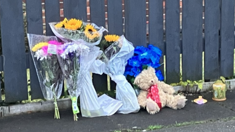 A teddy bear and bouquets of flowers are left by a fence in front of the house in Salford where two bodies were found on Monday