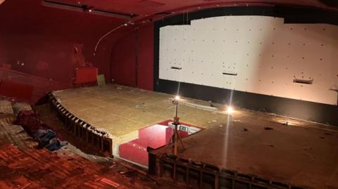 Inside of a cinema, where work is beginning to upgrade it. The screen has been removed and there is a white board in its place. There are floor boards where a stage used to be