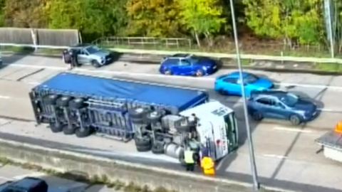 Aerial image of a blue and white lorry on its left side across a four-lane motorway. Four vehicles are parked to the left of the lorry