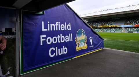 A view of a football stadium from the walkout tunnel. Linfield Football Club is written on a blue banner to the left of the tunnel wall against one of the stands. The football clubs crest is also on the banner.  