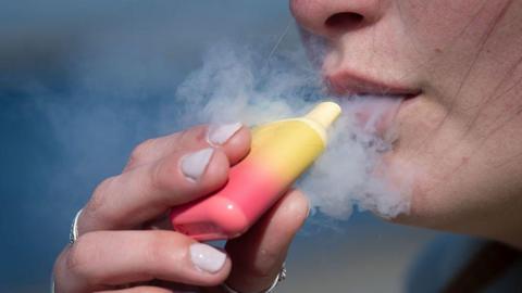 A woman poses for a photograph holding a pink and yellow single-use vape