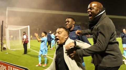 George Akhtar and Fabian Forde join celebrations with fans at Sixfields Stadium. They are running towards the away fans as red smoke from a pyrotechnic flare drifts over the goal behind them.