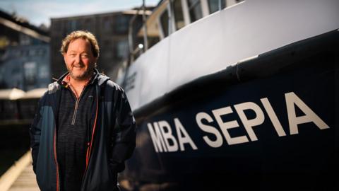 Dr Bryce Stewart wearing a rain coat and fleece is stood next to a yacht that has the lettering MBA Sepia on the side. 